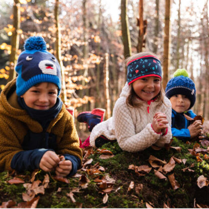 Drei Kinder beugen sich im herbstlichen Wald über einen Baustamm und lächeln in die Kamera